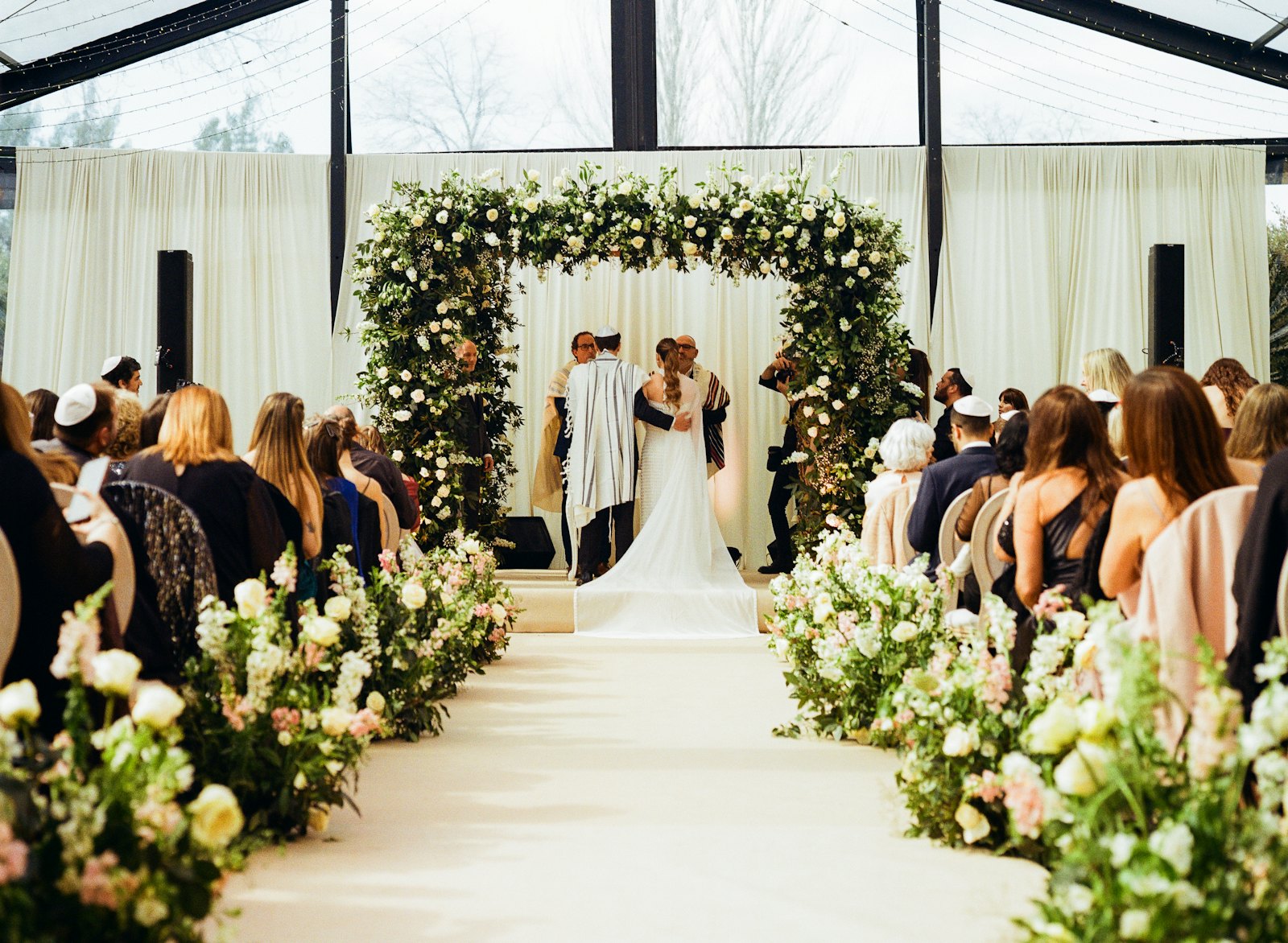 A couple under a floral chuppah with guests seated on both sides